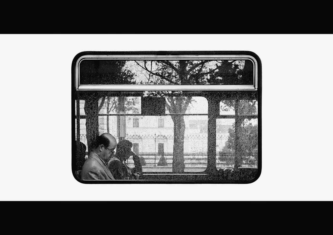 Black and white photo of people sitting on a bus or train, viewed through the window with rain droplets, trees, and buildings outside.