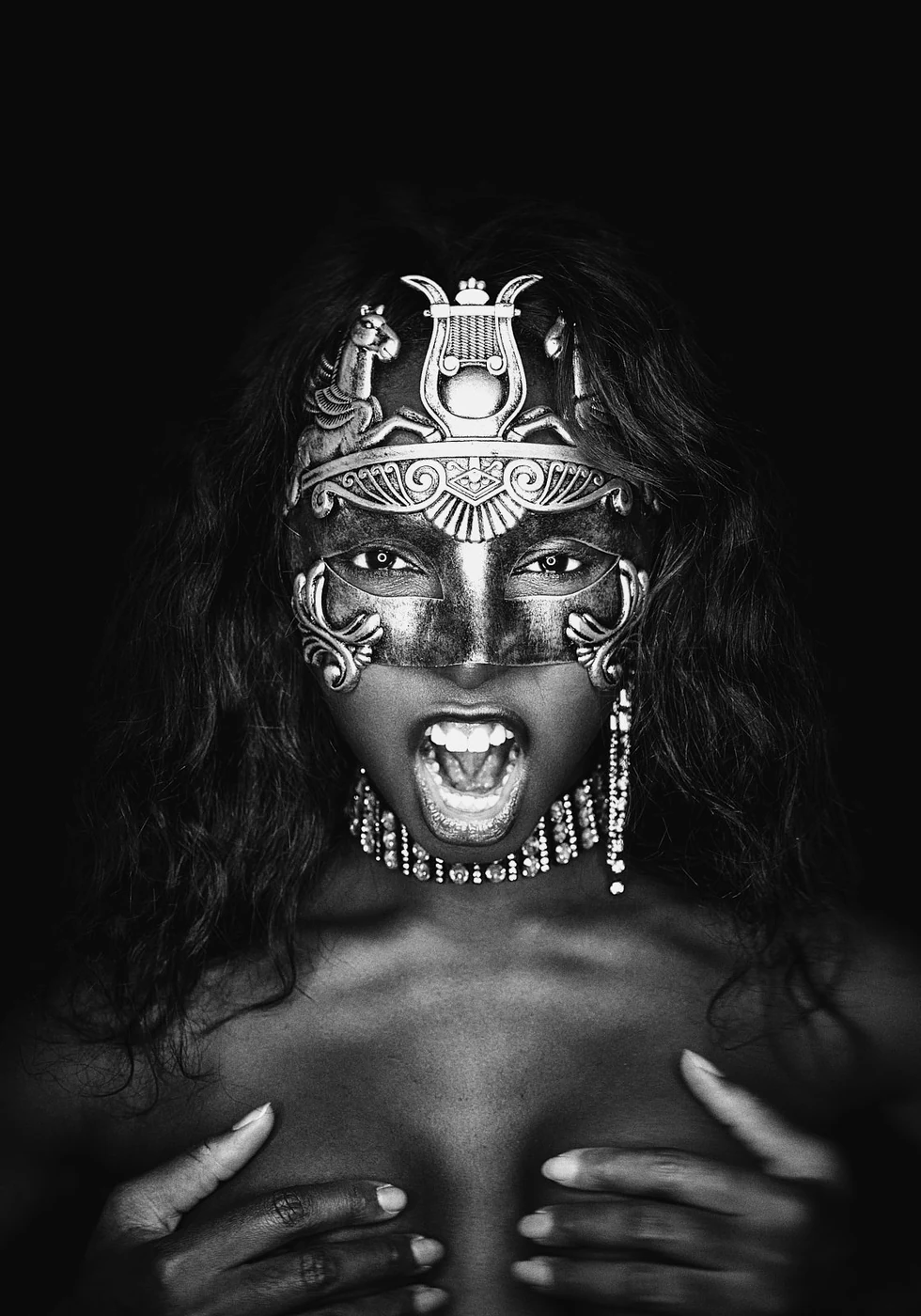 A woman wearing an ornate, tribal-style mask and jewelry, with long curly hair, shouting with her mouth open, holding her chest with both hands, against a black background.