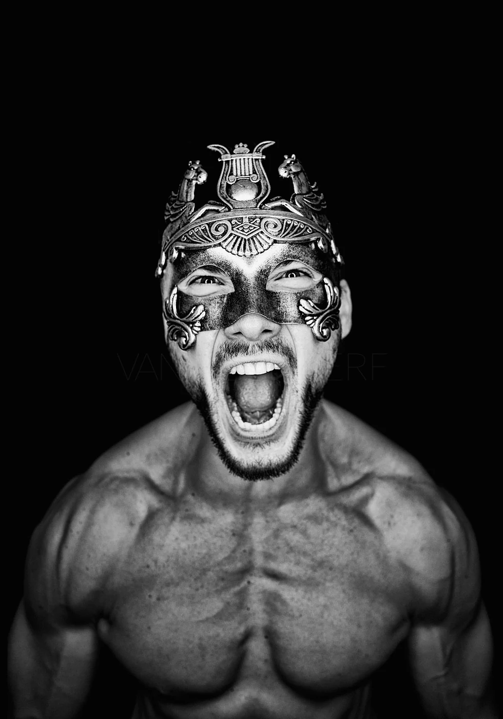 A shirtless man with a beard wearing an ornate metallic mask, shouting with his mouth open, against a black background.