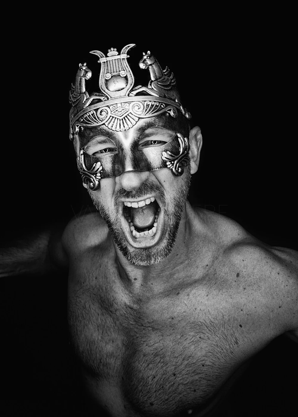 A shirtless man with a beard wearing an ornate mask and headpiece, shouting with his mouth open against a black background.