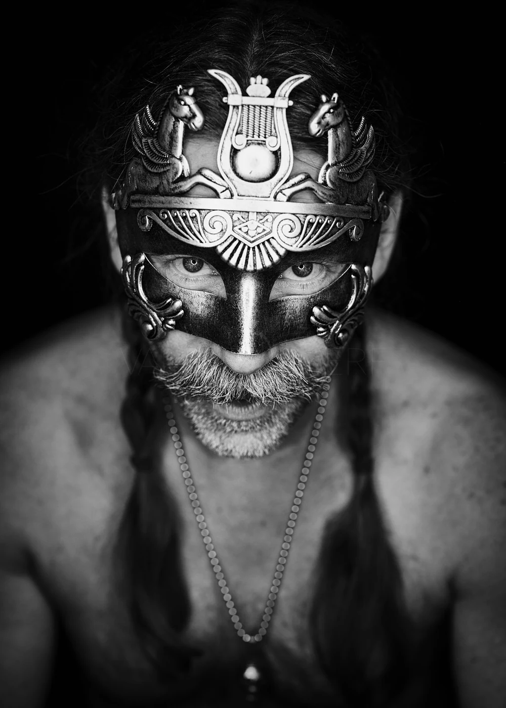 A man with long hair and a beard wearing a decorative mask that covers his eyes and forehead, with a dark background.