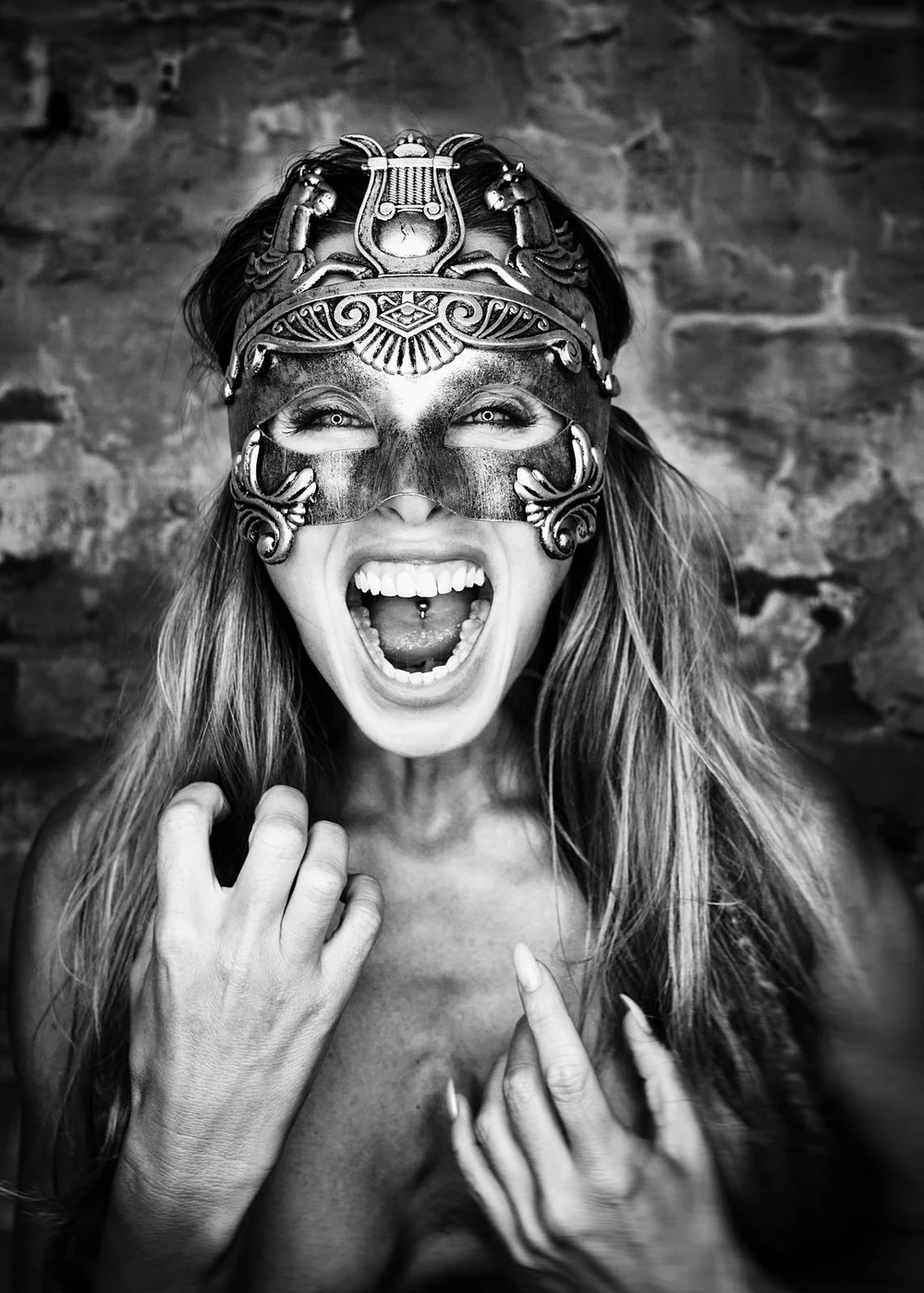 Woman wearing a decorative mask, smiling widely with open mouth, showing piercings, and holding her hair. Background is a brick wall.