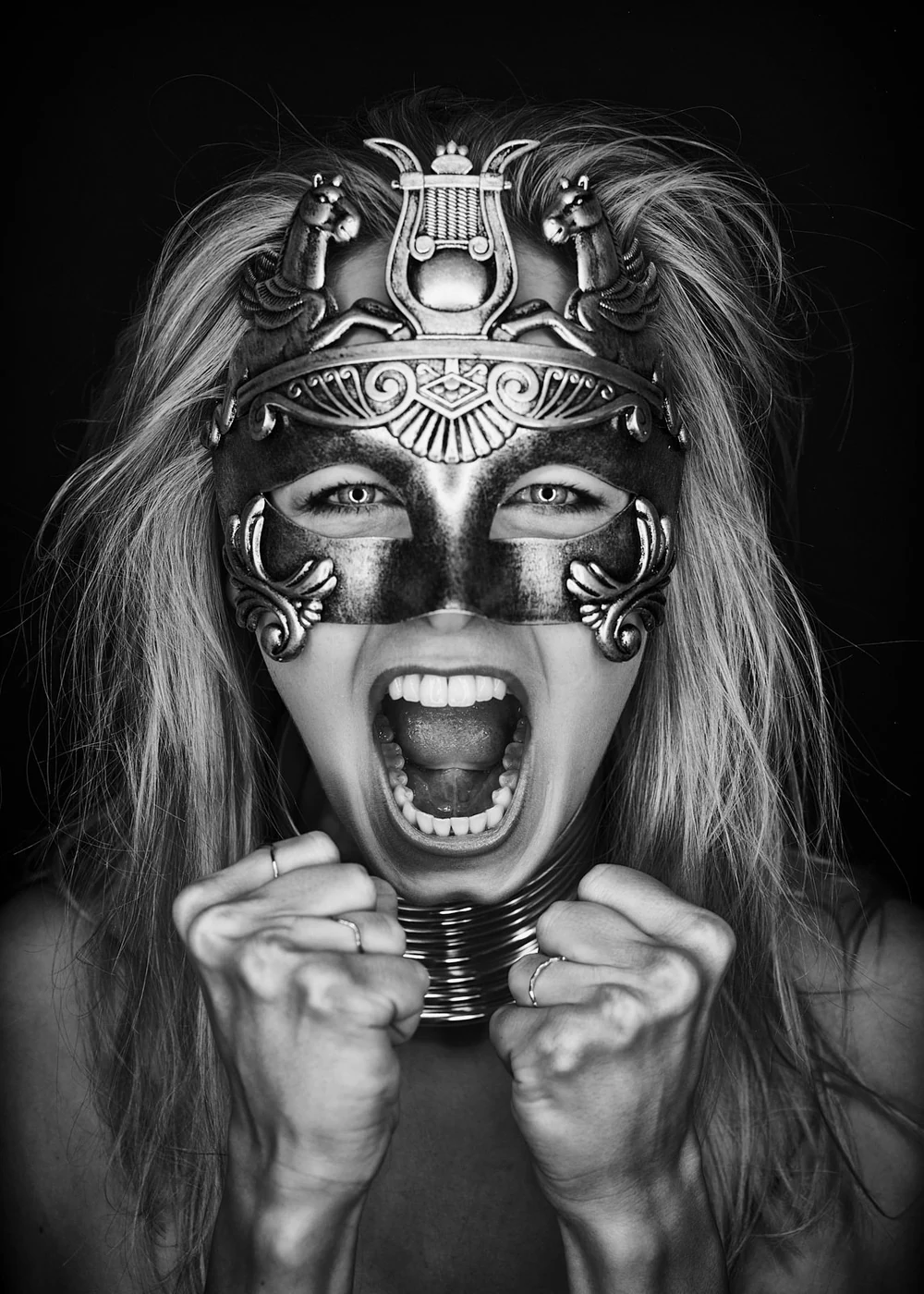 A woman with long hair wearing an intricate mask with animal and decorative designs, holding the mask at her chin, shouting with an open mouth, in black and white.