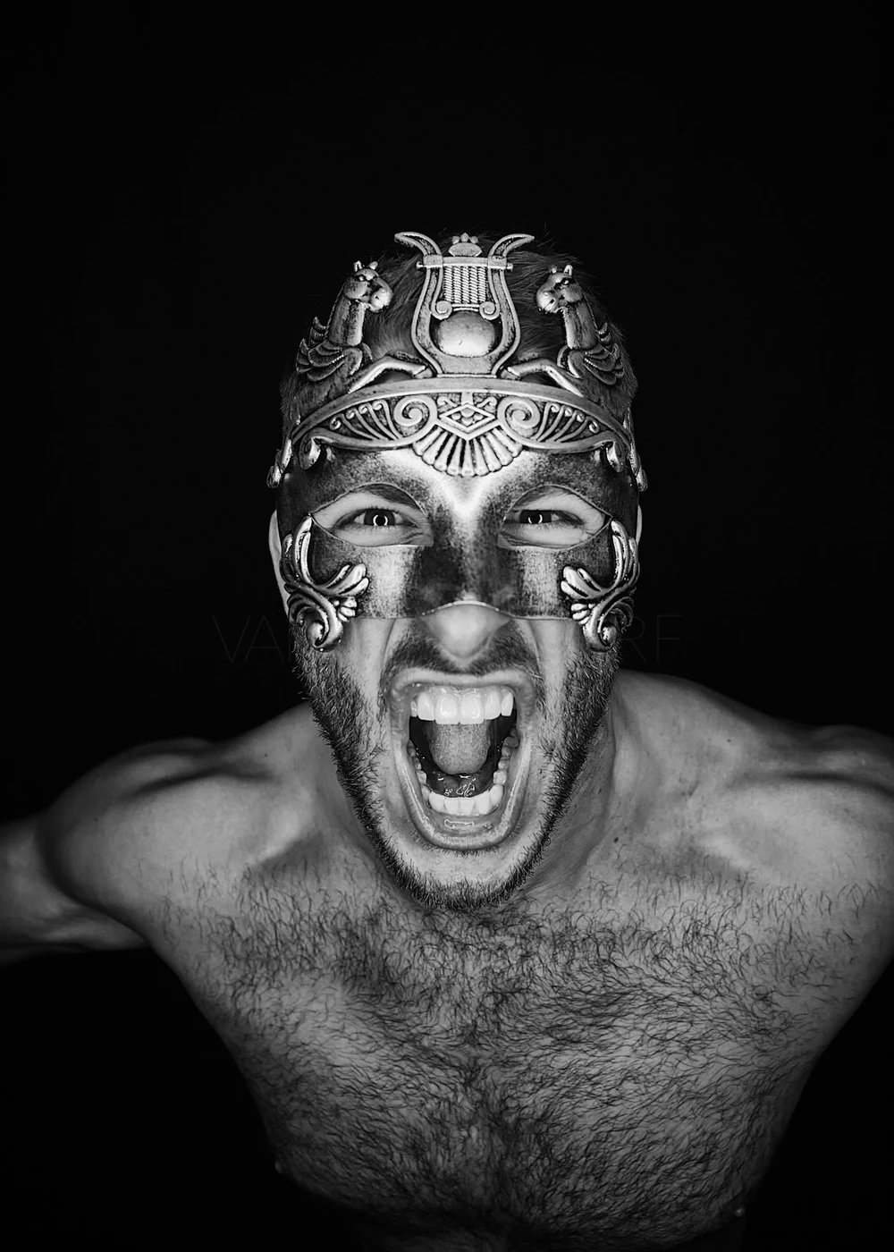 Black and white photo of a shirtless man with a beard wearing an ornate mask, screaming with his mouth open and eyes wide, against a black background.