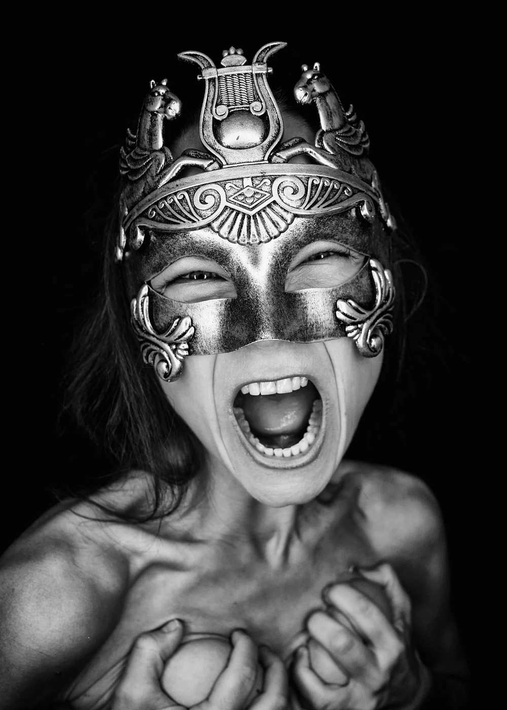 A woman wearing an ornate metallic mask with decorative elements, including horse figures and a lyre. She is shouting with her mouth wide open, and her hands are gripping her chest. The image is in black and white.