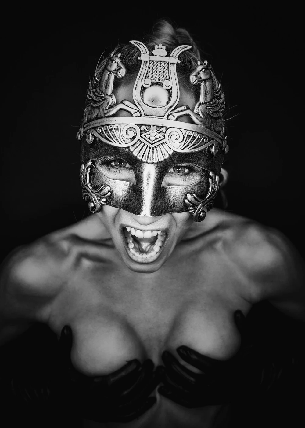 A person wearing a decorative mask with an intricate design and horse figures, expressing a fierce and intense emotion, in black and white photography.