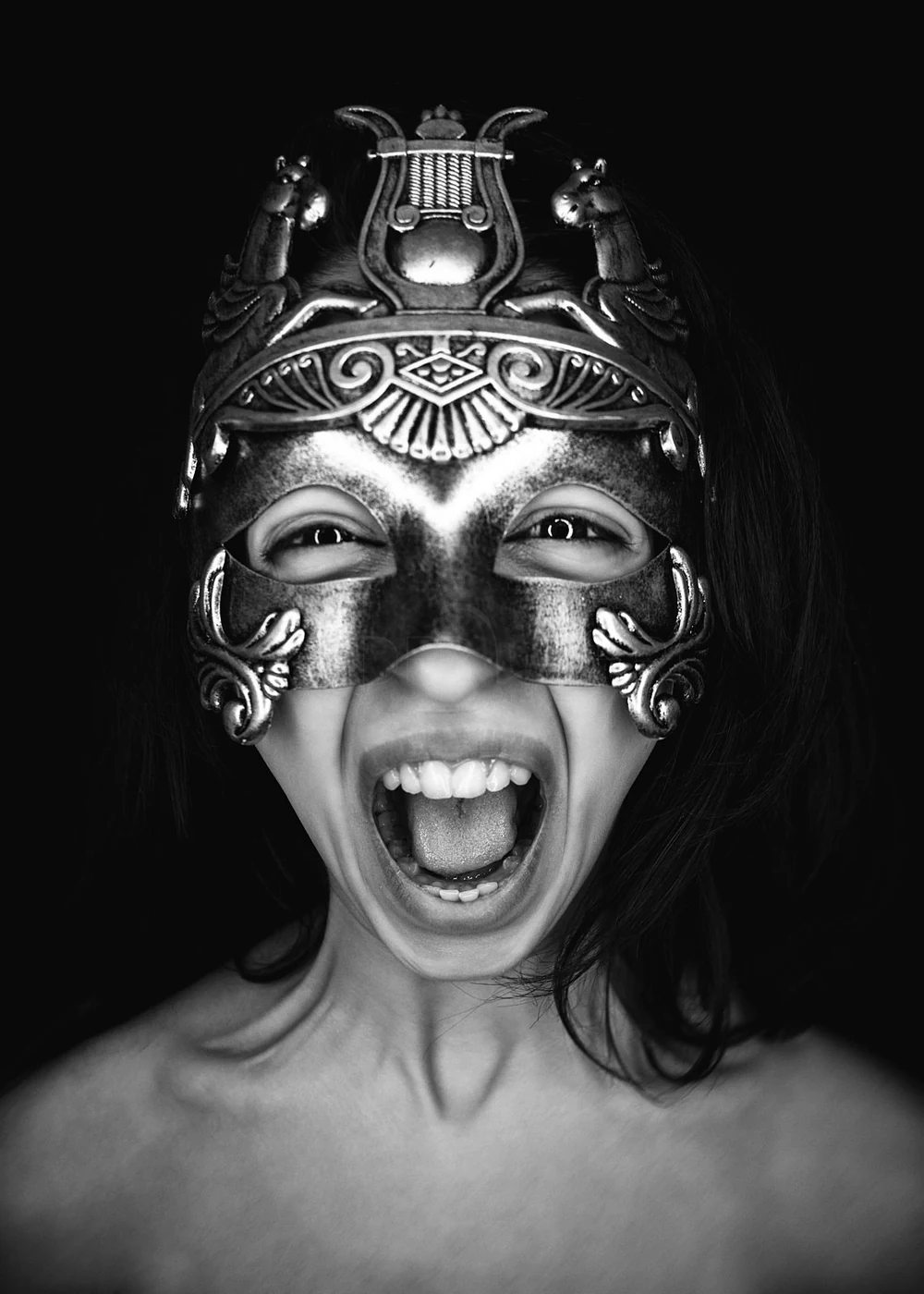 A person wearing an ornate, metallic mask with animal and decorative elements, smiling with mouth open and tongue out, set against a black background.