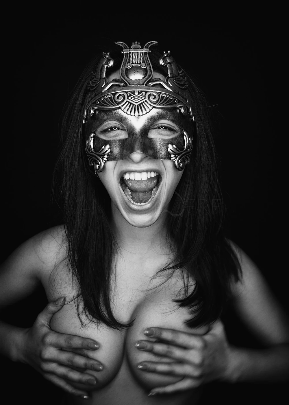 A woman wearing an ornate mask, smiling broadly, with her hands covering her breasts.