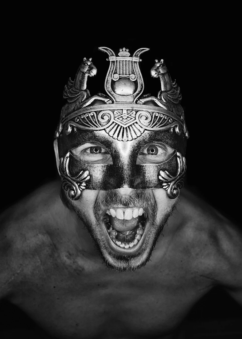 Black and white photo of a shirtless man wearing an ornate, metallic mask with decorative elements and animal figures. The man is shouting or screaming with his mouth open wide, showing teeth, and looking directly at the camera.