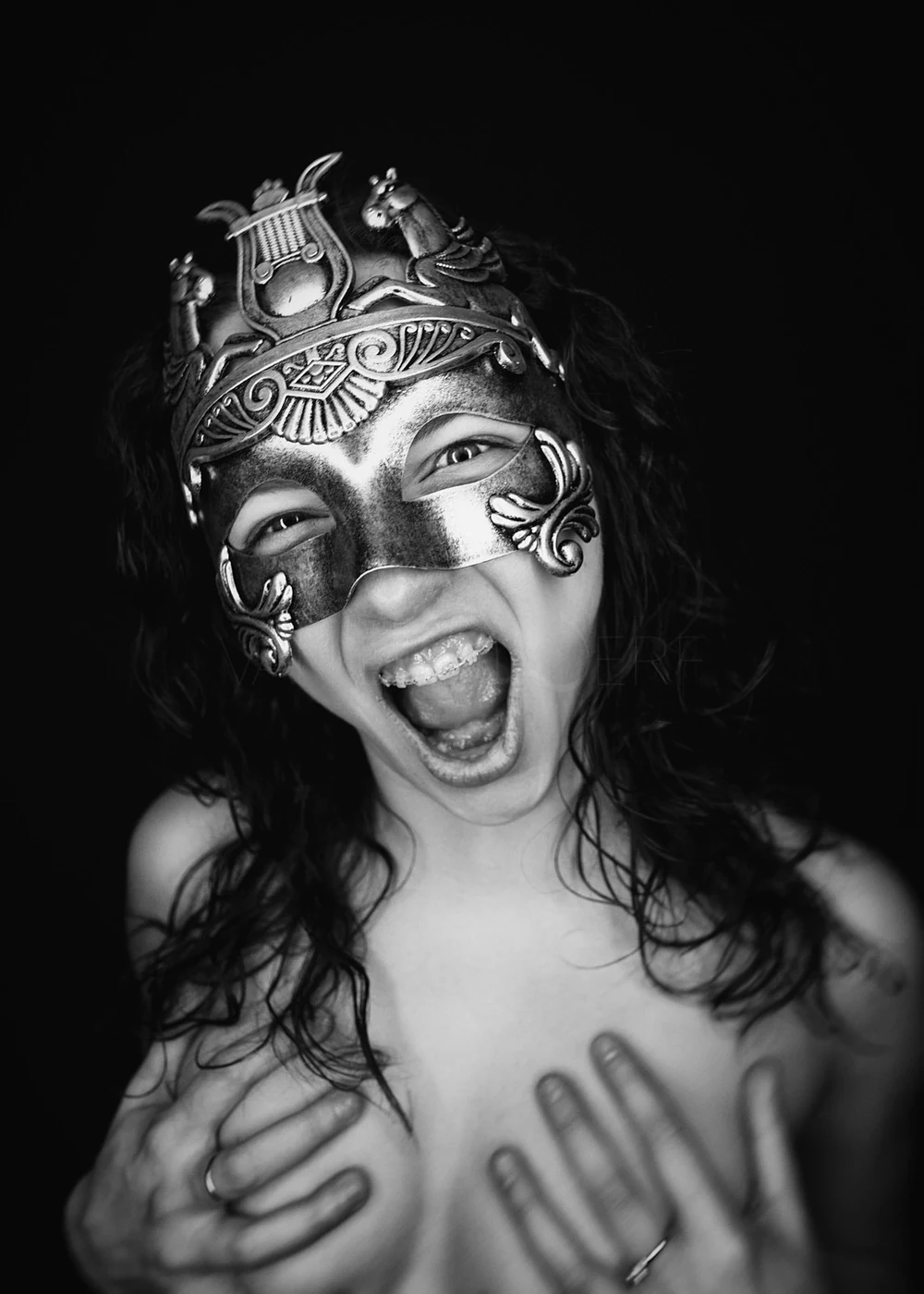 A woman with long, curly hair wearing an ornate mask, smiling and holding her chest with hands, against a black background.
