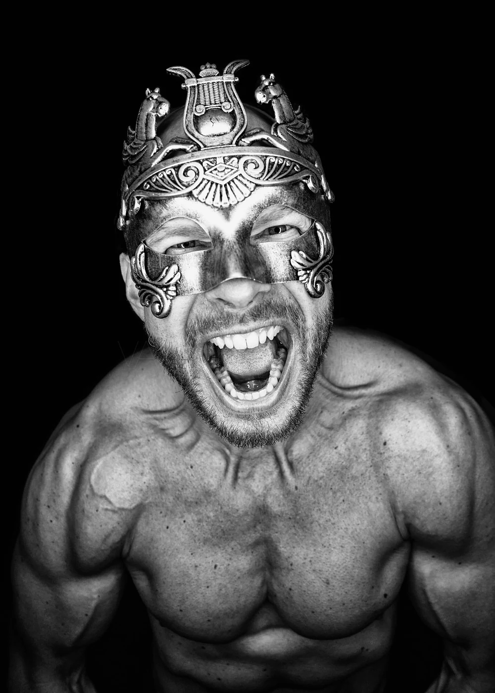 A muscular man with a beard, wearing a decorative metallic mask and an ornate headdress, shouting with an open mouth against a black background.