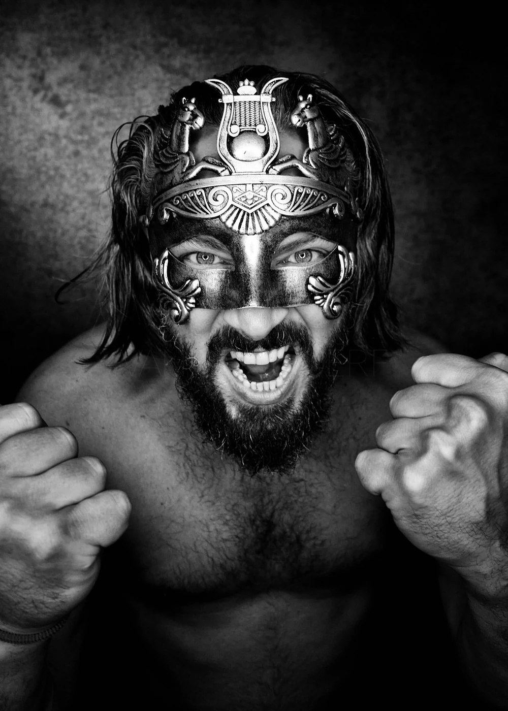 A muscular man with long hair and a beard wearing an ornate mask, showing his clenched fists and shouting fiercely.