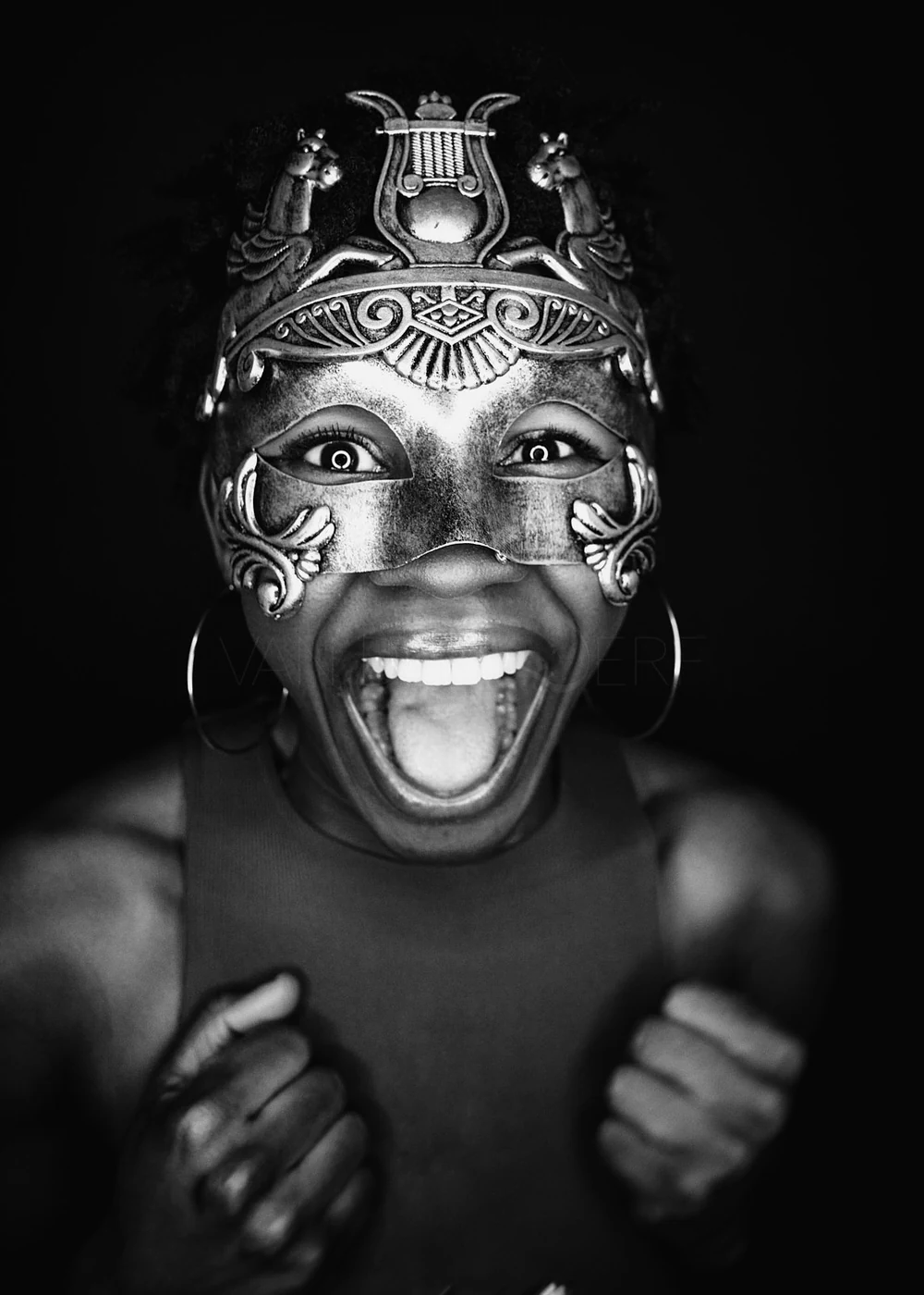 A woman wearing a decorative mask with an open-mouthed smile, showing her teeth, with clenched fists near her chest, in black and white.