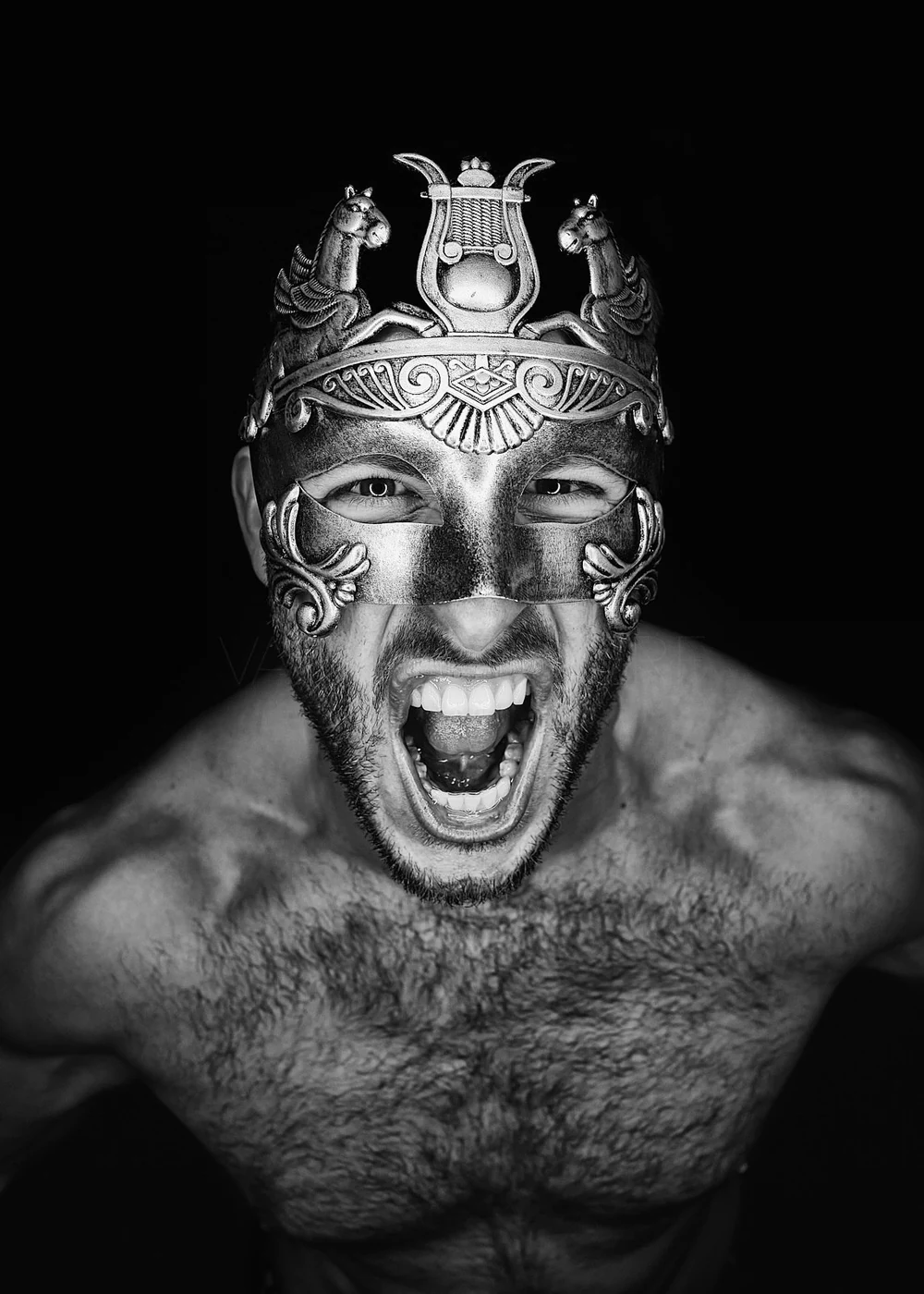 A shirtless man wearing a metal mask and an ornate headpiece with two horse figures, shouting or roaring with an intense expression.