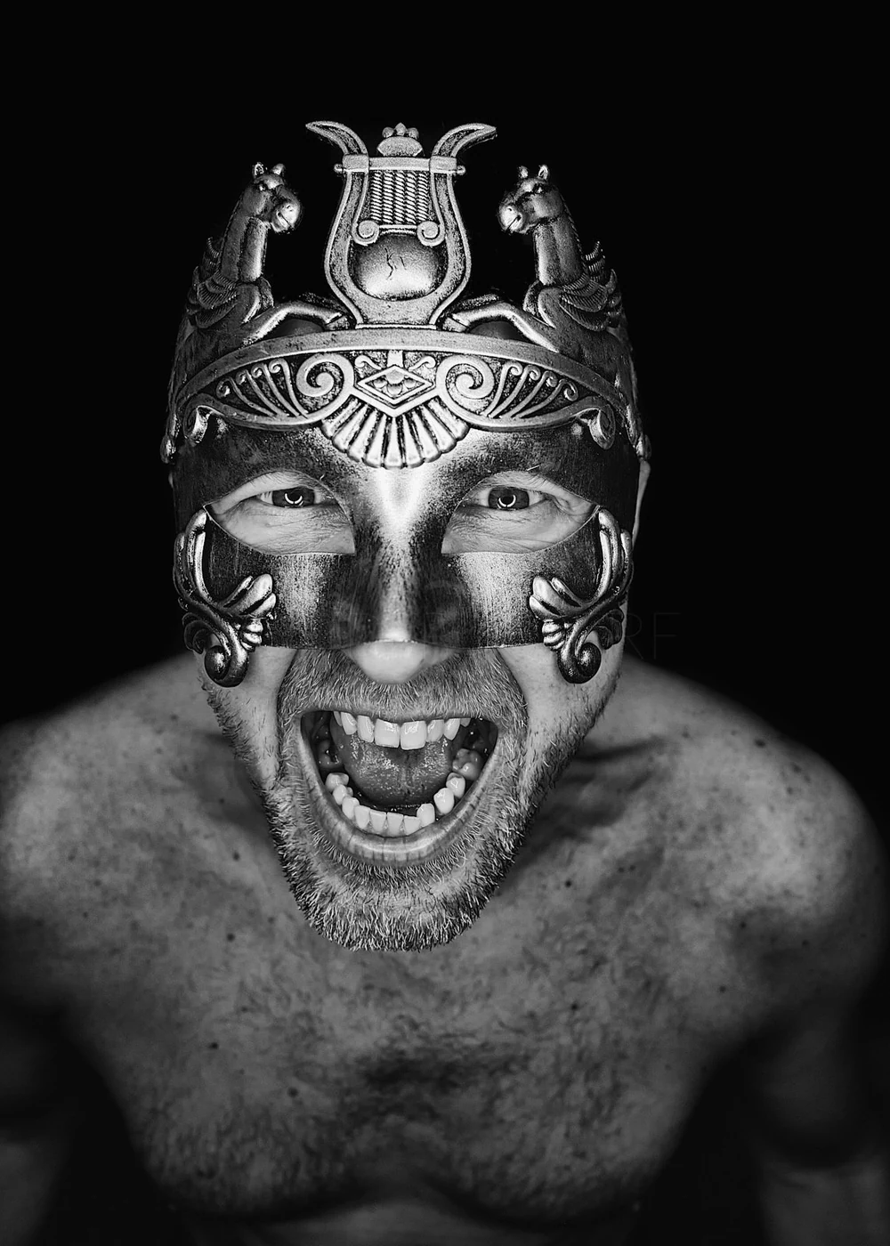 A shirtless man wearing an ornate, decorative mask with a warrior or tribal design, showing an excited or fierce expression against a black background.