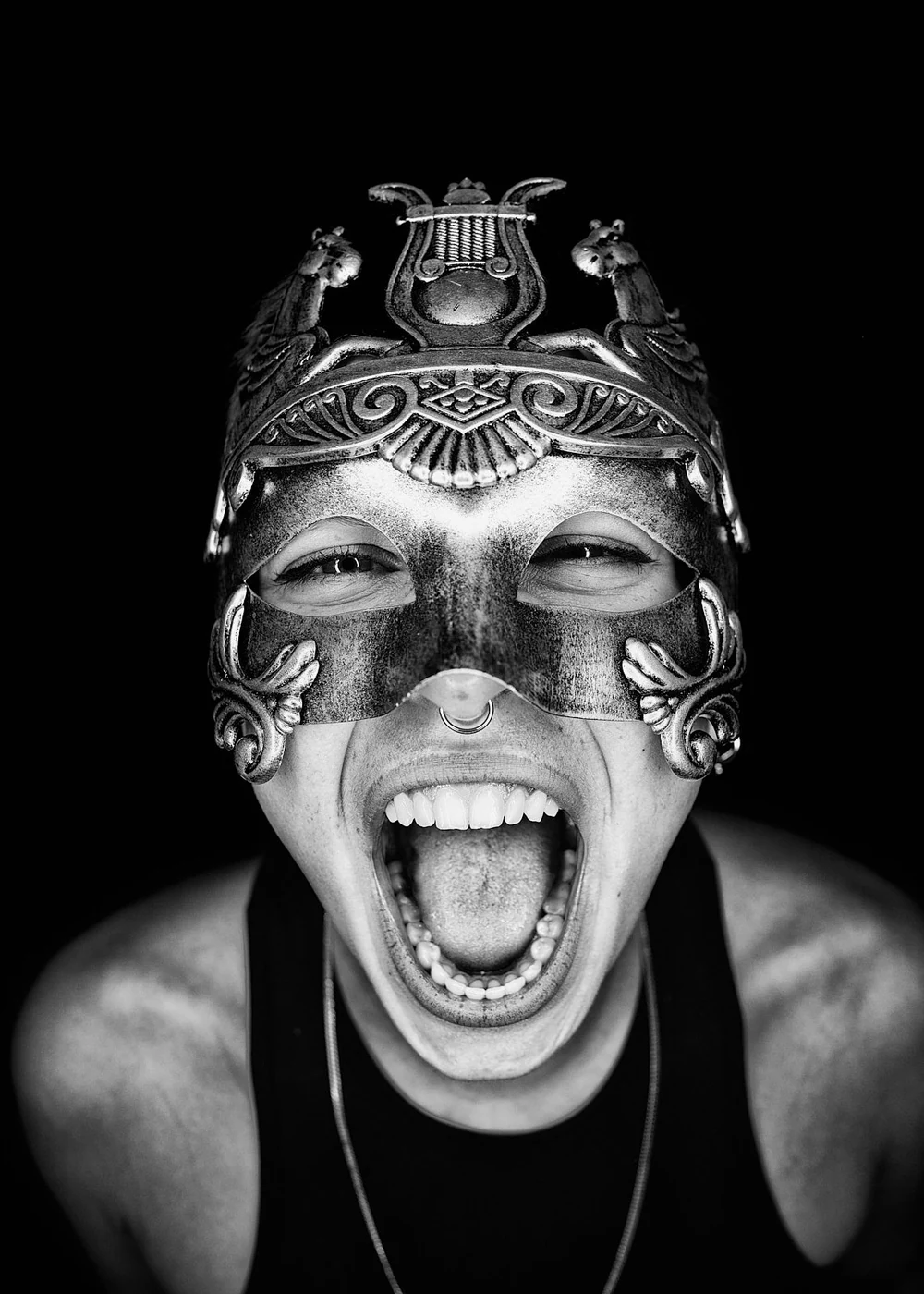 A person wearing an ornate mask with mythical creatures, shouting with mouth wide open, black background.
