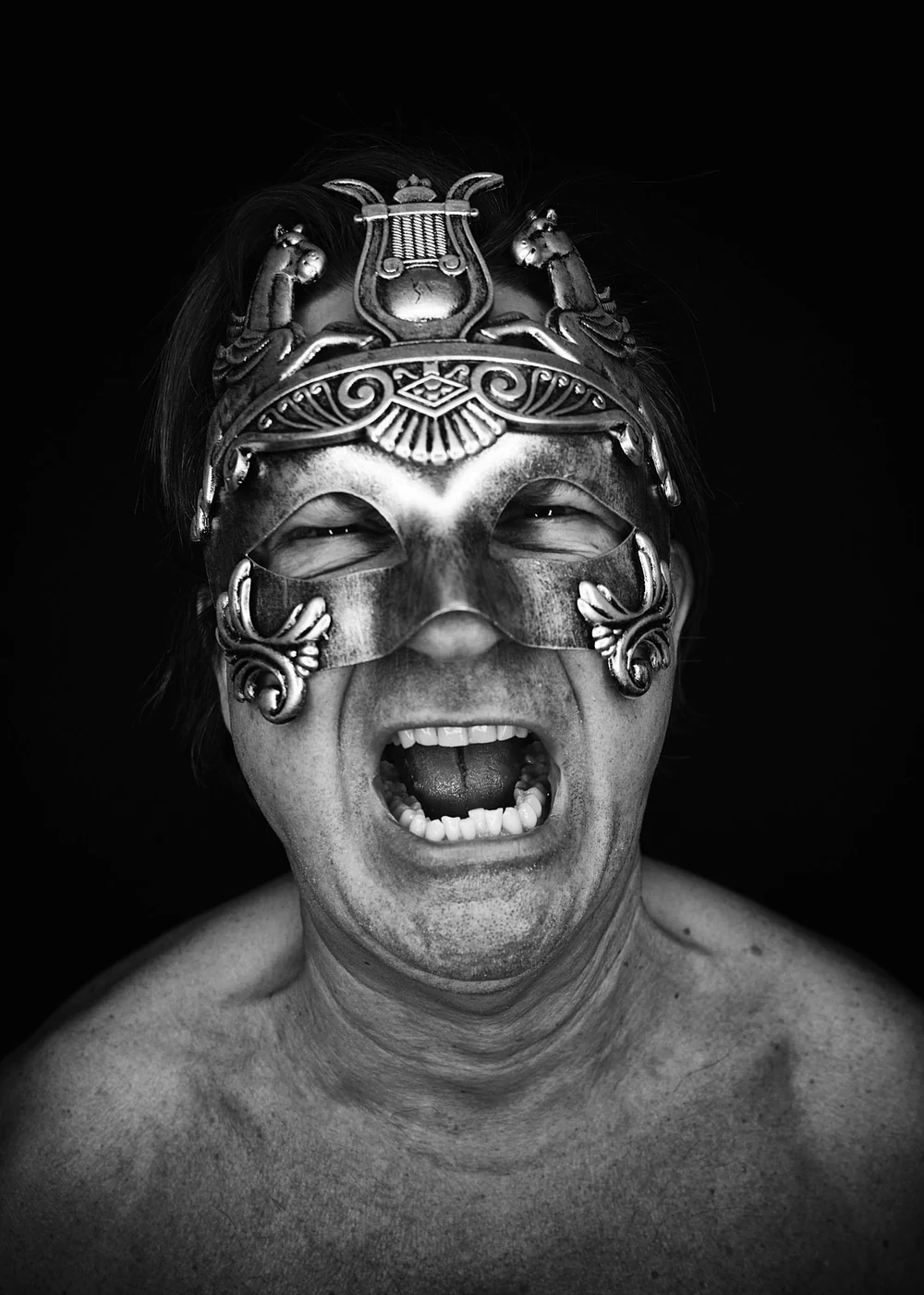 A shirtless man wearing an ornate metallic mask and a decorative headpiece, with a wide open mouth as if shouting or laughing, against a black background.