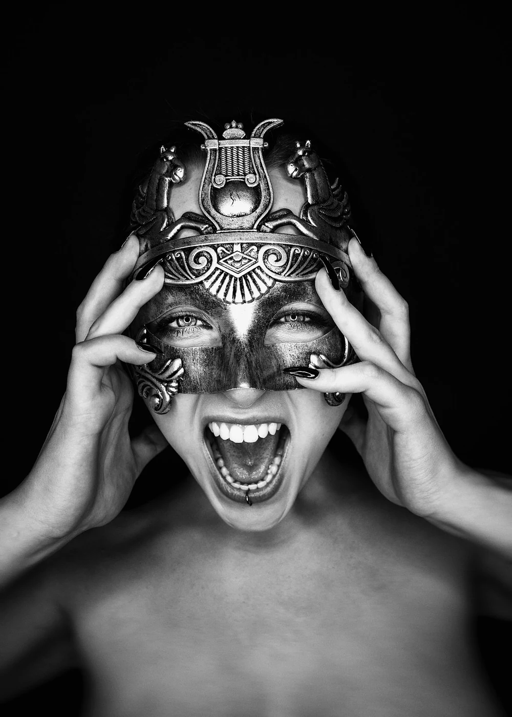 A woman with a pierced lip holding an ornate mask with both hands, revealing her face and mouth, against a dark background.