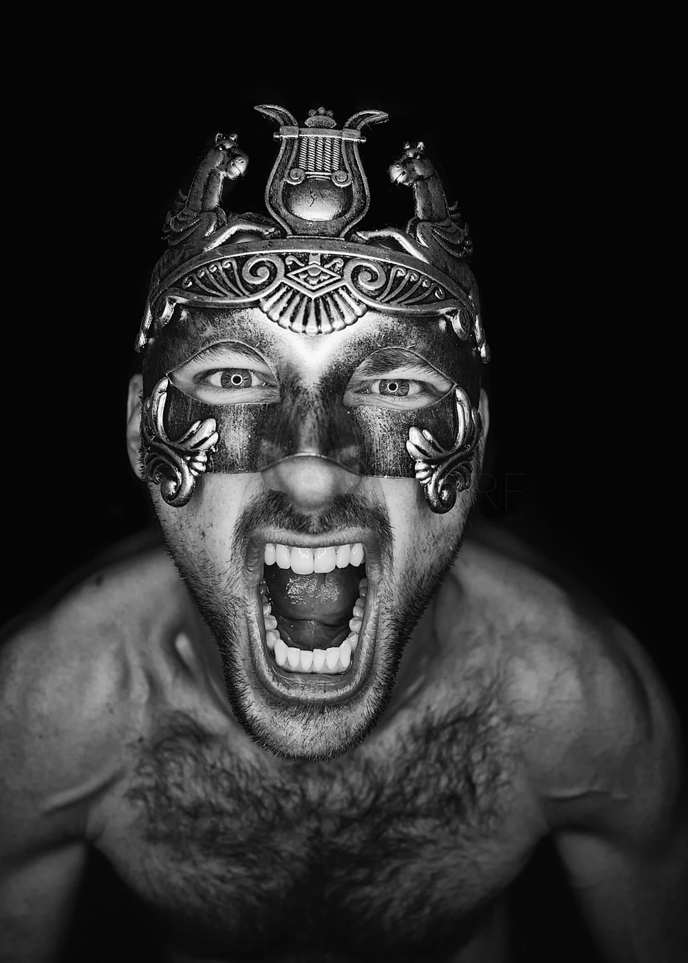 A shirtless man with a beard wearing an ornate mask with decorative elements and animal figures, shouting with his mouth open, against a black background.
