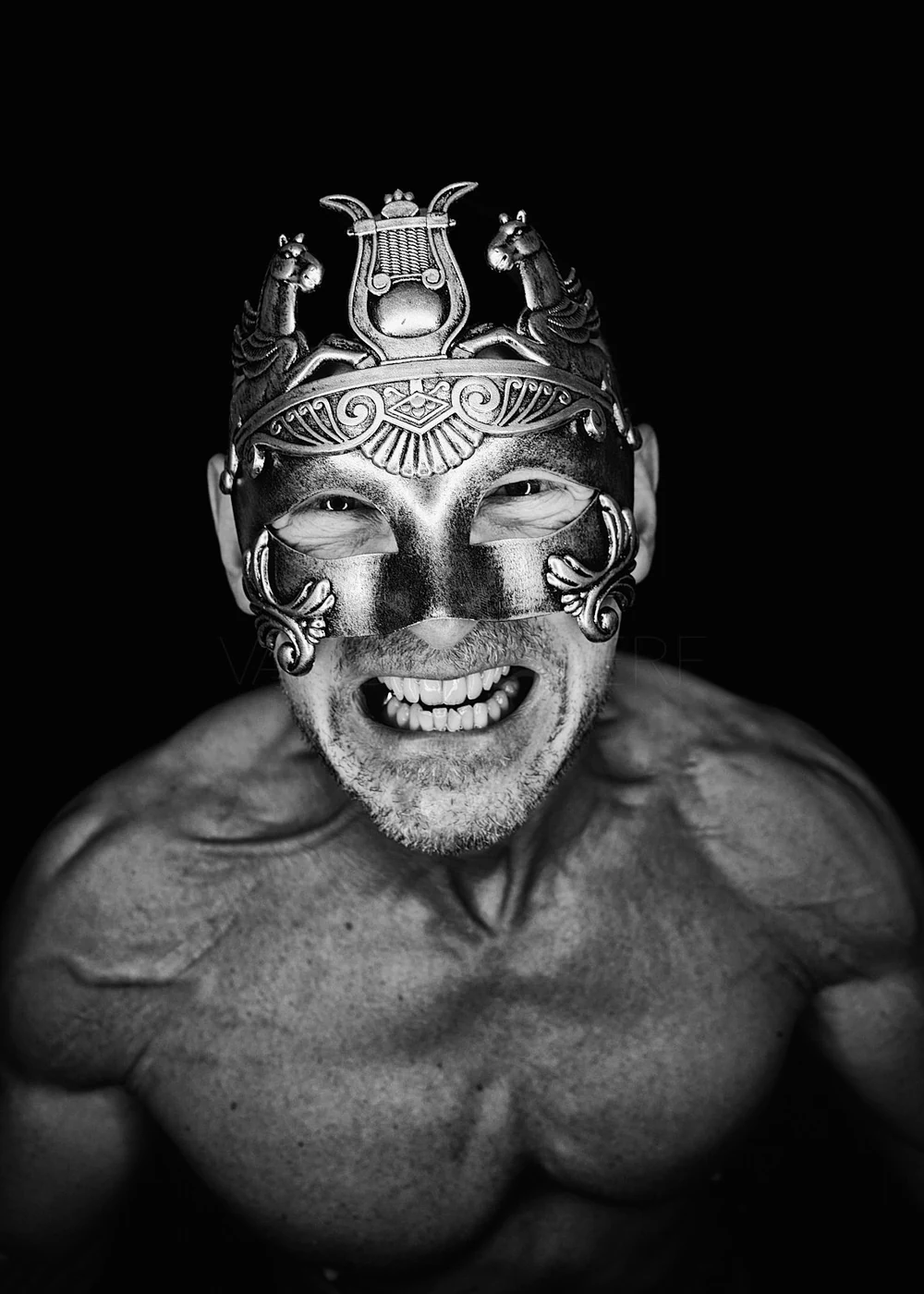 A muscular man smiling while wearing an ornate, decorative mask and a detailed, elaborate headpiece with horses and other intricate designs, set against a black background.