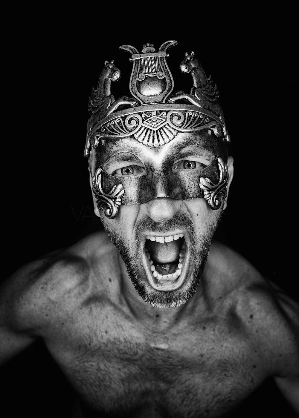 A shirtless man with a beard wearing a decorative metal mask with intricate patterns and two small horse figures on top, shouting with his mouth open against a black background.