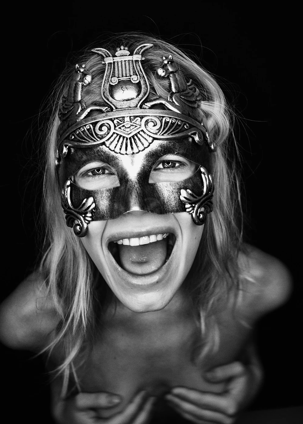A person with long hair wearing a decorative mask, smiling widely and showing open mouth, with a dark background.