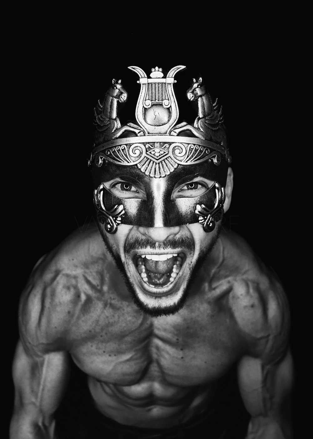 A shirtless man wearing an ornate, decorative mask with a headdress featuring animals and patterns, and expressing a fierce, shouting emotion. The photo is in black and white.
