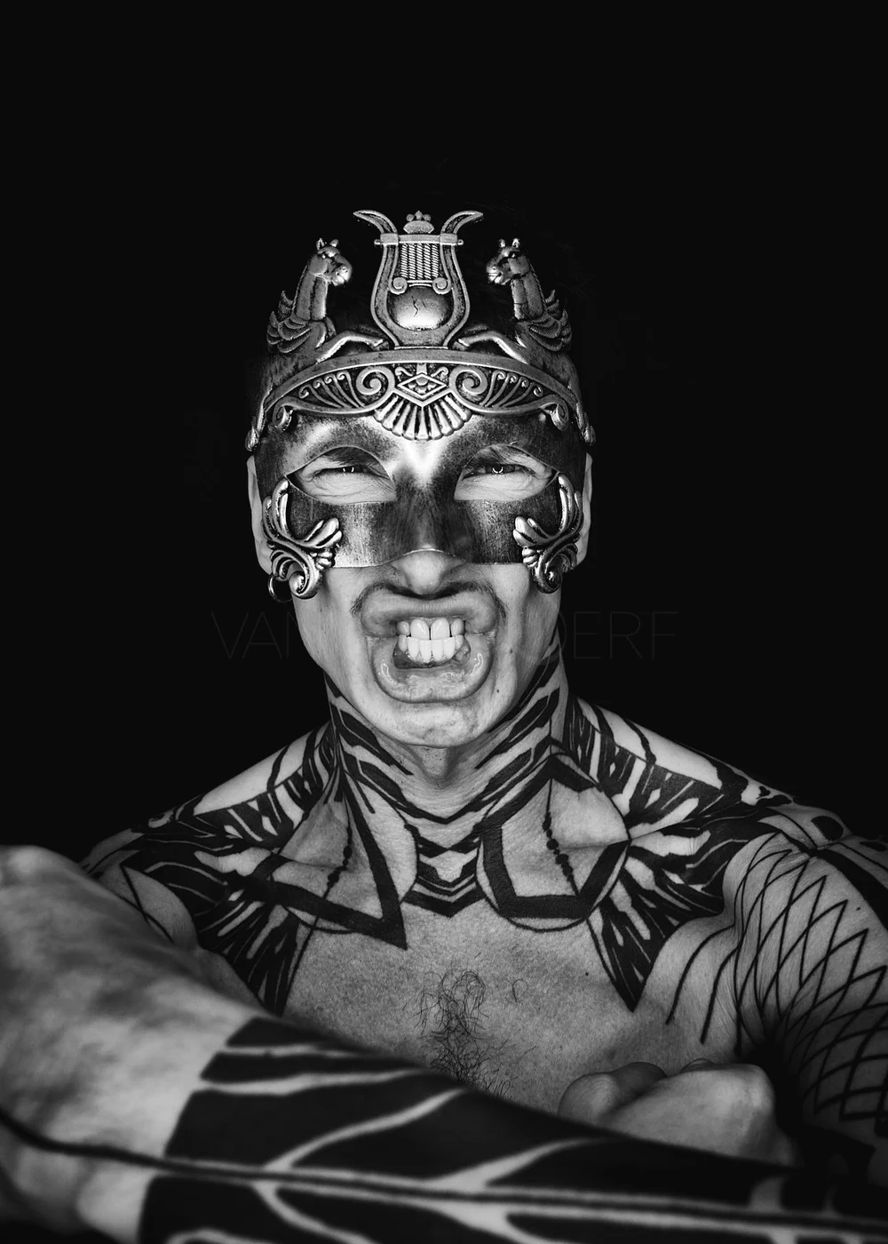 A person wearing a decorative tribal mask with metallic details, showing an aggressive facial expression and tattoos on their chest and arms, against a black background.