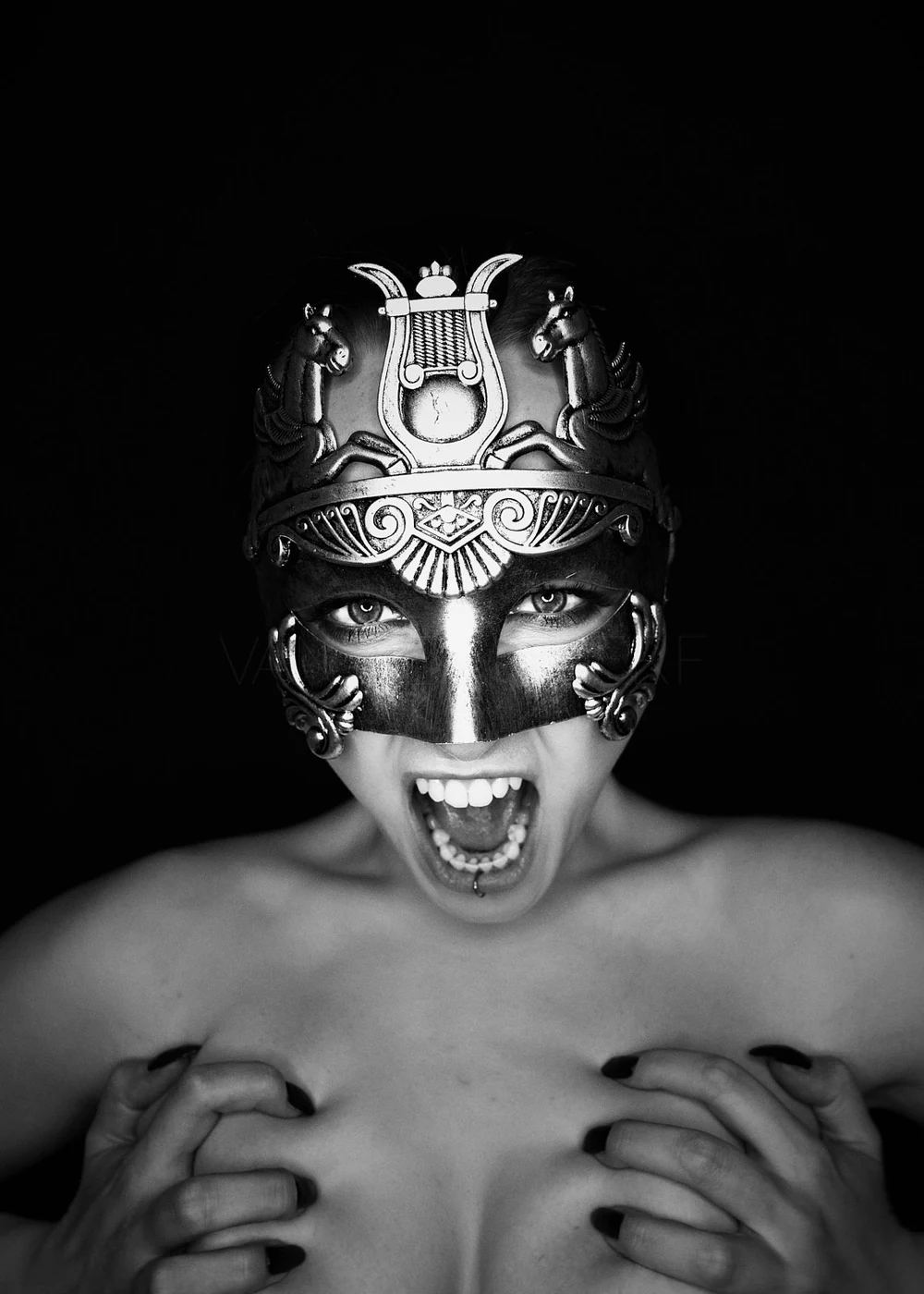 Black and white photo of a woman wearing an ornate metallic mask, shouting with her mouth open, revealing her teeth and tongue piercings. She has dark nail polish and is holding her breasts with her hands. The background is black.