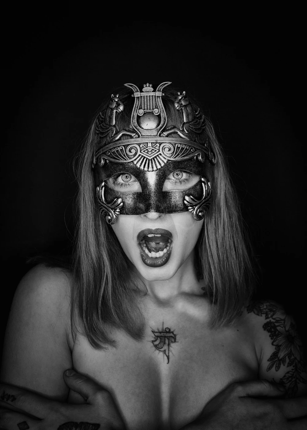 A woman wearing an ornate metallic mask covering her eyes and forehead, with an open mouth expressing surprise or shouting, long hair, and tattoos on her shoulders and chest, set against a dark background.