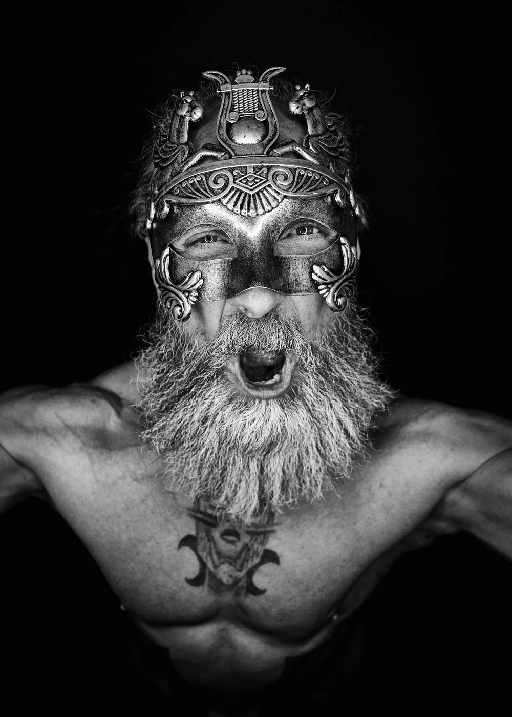 A shirtless man with a long beard wearing a metallic, ornate mask covering part of his face, with an open mouth and intense expression, black background.