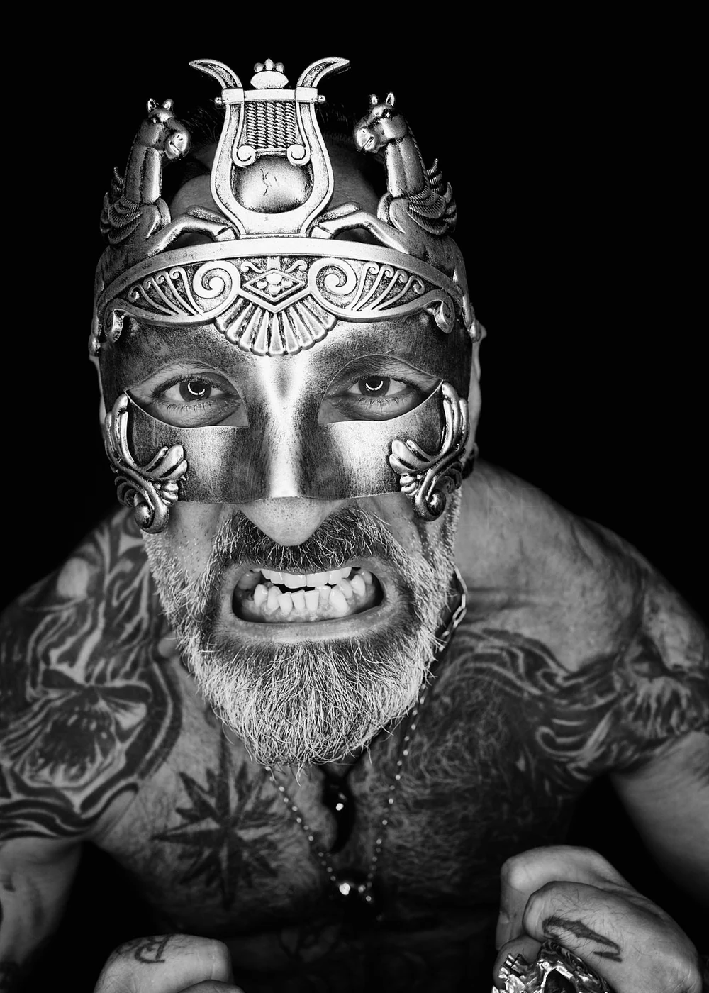 A man with a beard wearing an elaborate metal mask with horse and mythological creature designs, showing his intense eyes and gritted teeth, tattoos, and jewelry, against a black background.