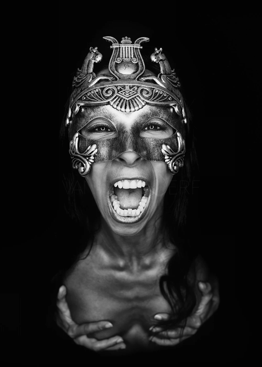 A woman wearing an ornate mask with animals and musical symbols, with her mouth open in a scream, holding her chest with her hands.
