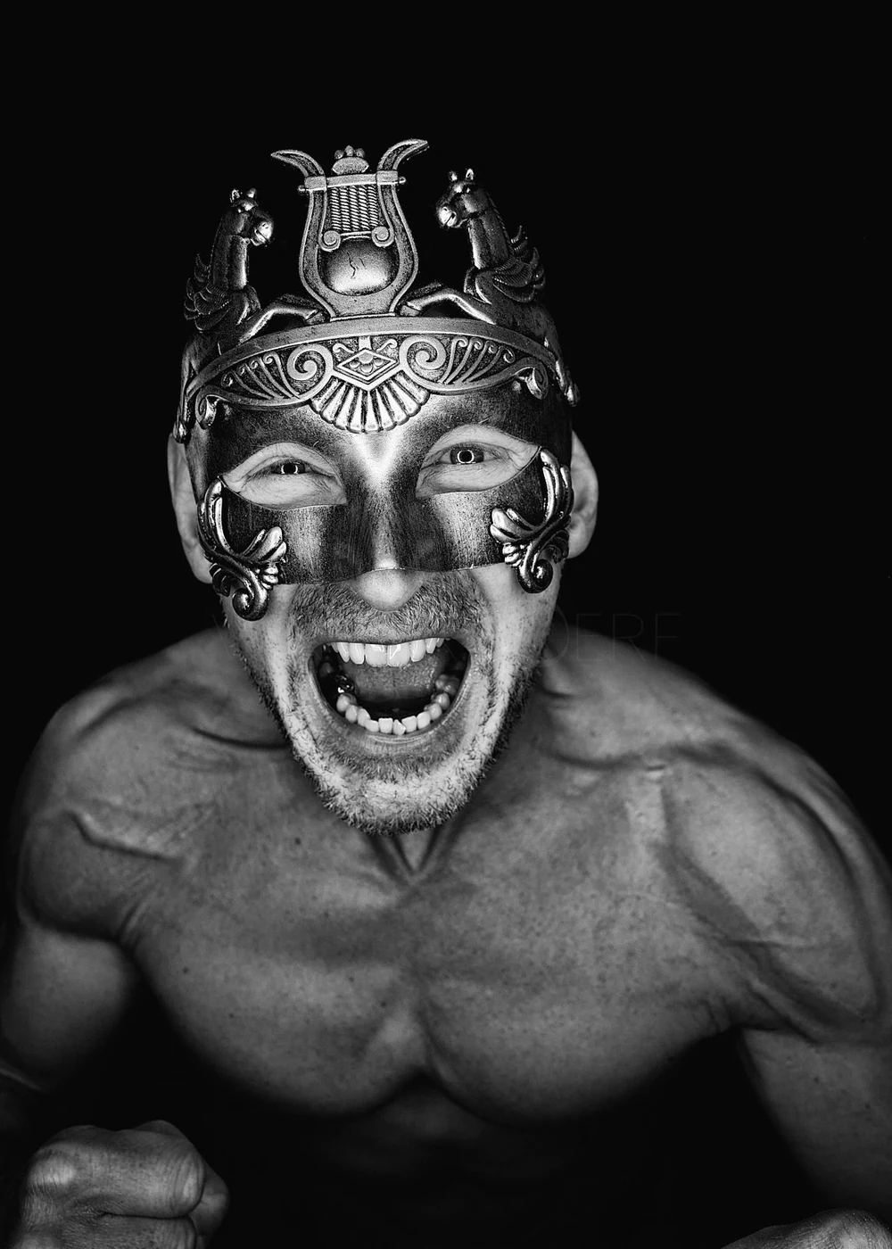 A shirtless man with a beard wearing an ornate, face-covering mask that resembles a Roman or Greek style with decorative elements and two horses on top. The man is smiling broadly, showing his teeth, and appears to be shouting or cheering, with his f