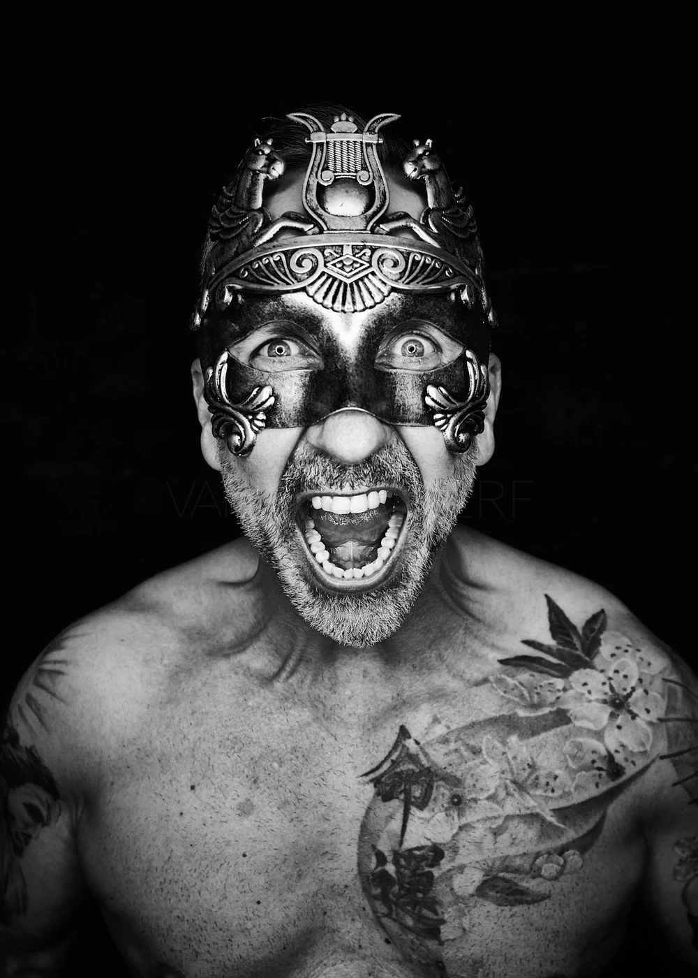 Black and white photo of a shirtless man wearing a decorative mask, showing his teeth and shouting, with tattoos on his chest and arm.