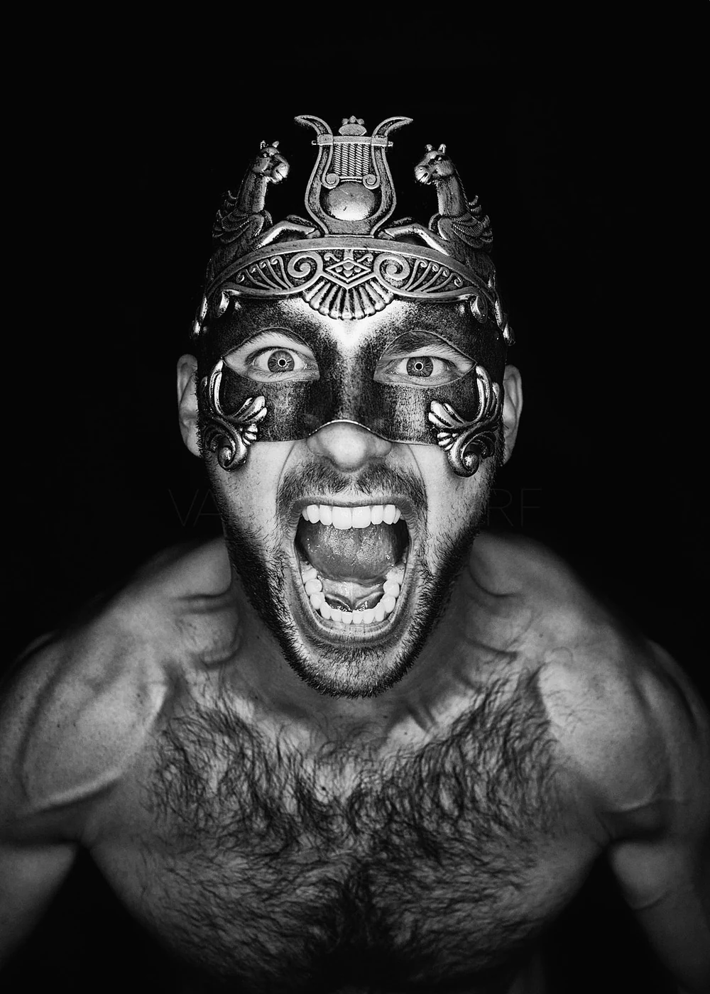 A shirtless man wearing a decorative metal mask with an open mouth, showing teeth, and looking directly at the camera against a black background.