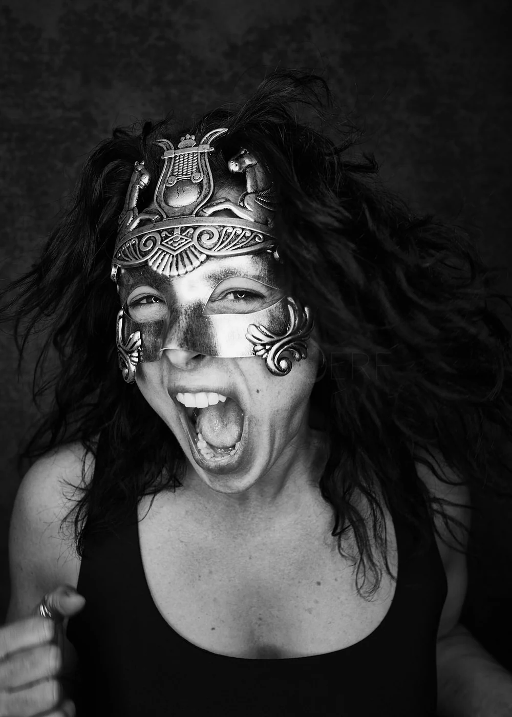 A woman with wild hair wearing an ornate metallic mask and smiling broadly, showing her teeth and tongue, in a black and white photo.