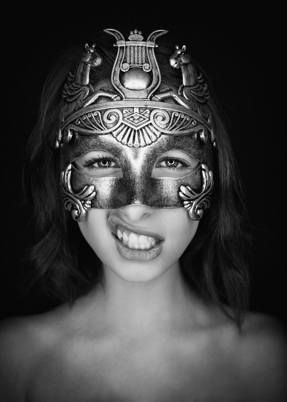 A woman with medium-length hair wears an ornate metallic mask covering her eyes and upper face, with intricate designs and protruding decorative elements. She is smiling with her teeth showing and her tongue slightly out, against a black background.