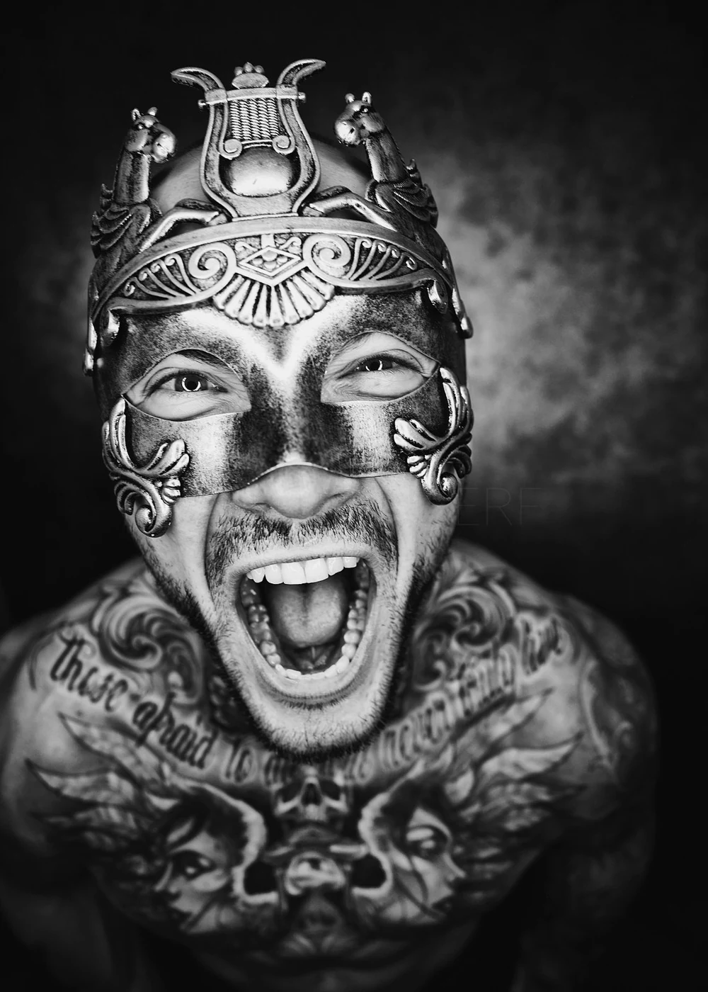 A man wearing a decorative mask with animal and tribal motifs, shouting with his mouth open, showing tattoos on his chest and neck.