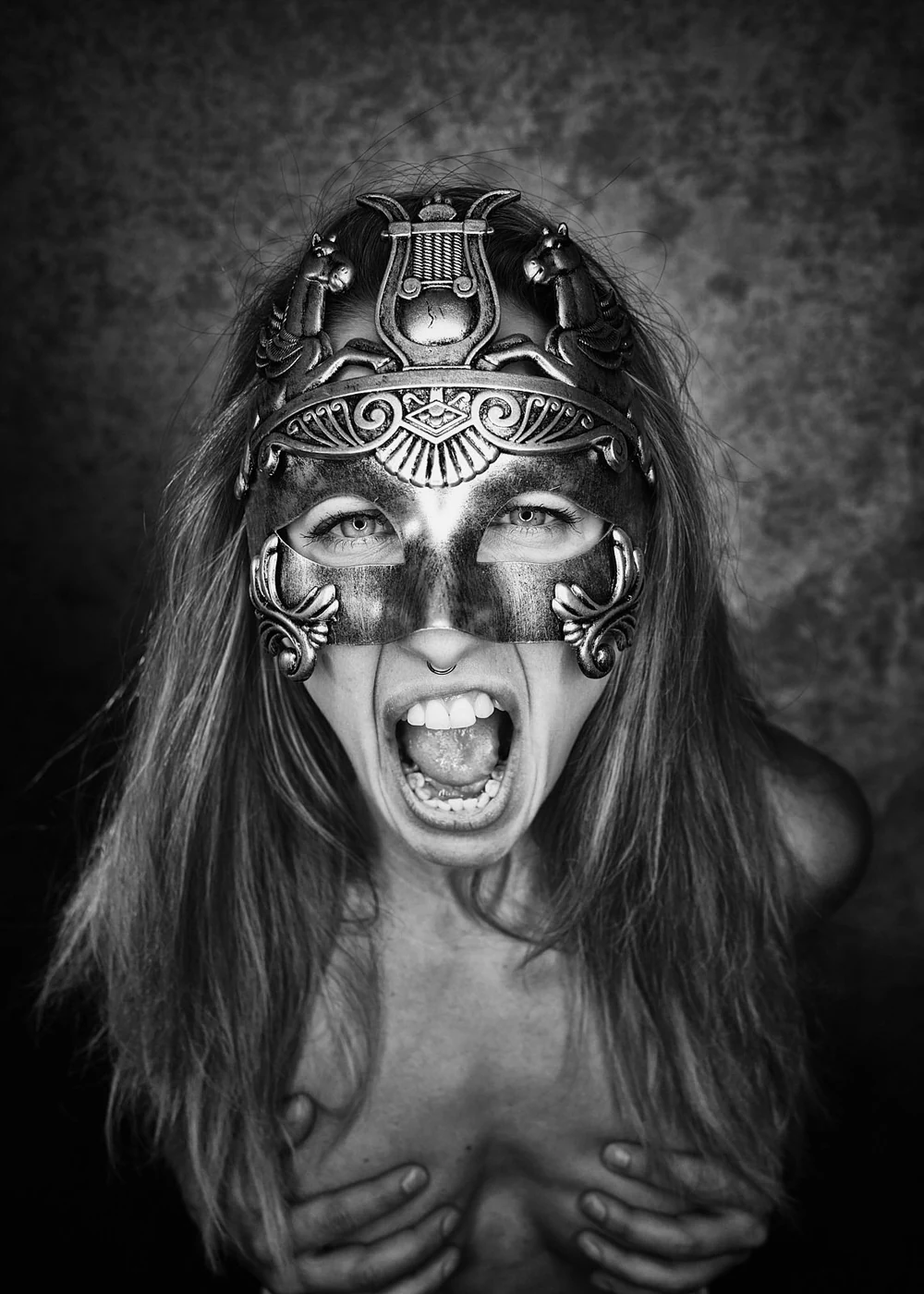 A woman with long hair wearing an ornate metal mask with Egyptian motifs and animal figures, yelling with an open mouth, holding her breasts.