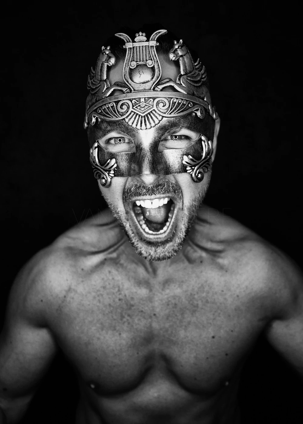 Black and white photo of a shirtless man wearing an ornate silver mask with a fierce expression, shouting with mouth open wide, against a dark background.