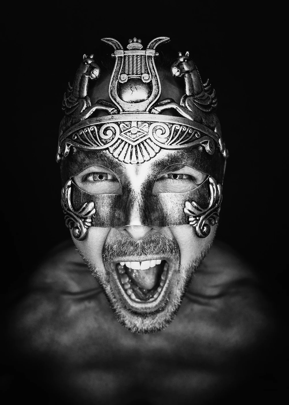 A man with a beard wearing a metal mask and an ornate crown, shouting with his mouth open, depicted in black and white.