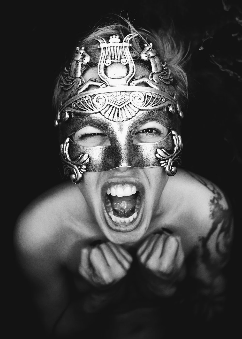 A person wearing a decorative mask with animal motifs, showing an aggressive facial expression with mouth open and fists clenched, against a dark background.