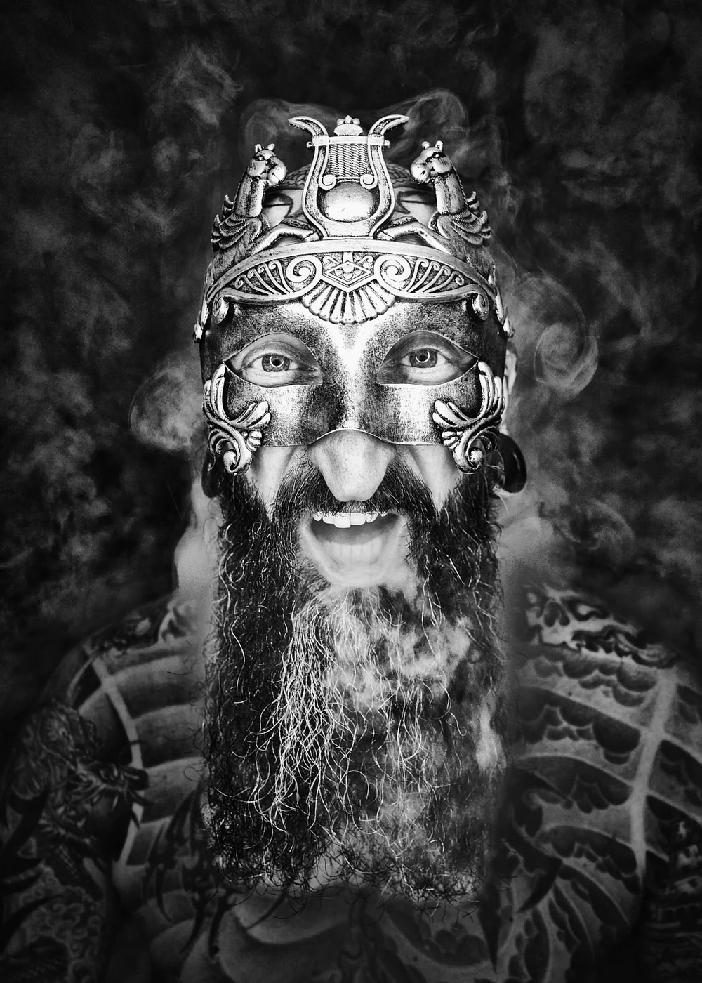 A man with tattoos on his shoulders and chest, wearing a decorative metallic mask and an ornate headdress with animal figures. He has a long beard, and smoke surrounds him. His eyes are visible through the mask.