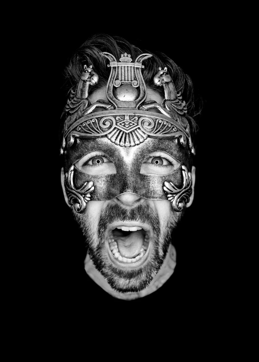 A man with a beard wearing an ornate mask featuring a scorpion design, shouting with an open mouth, against a black background.