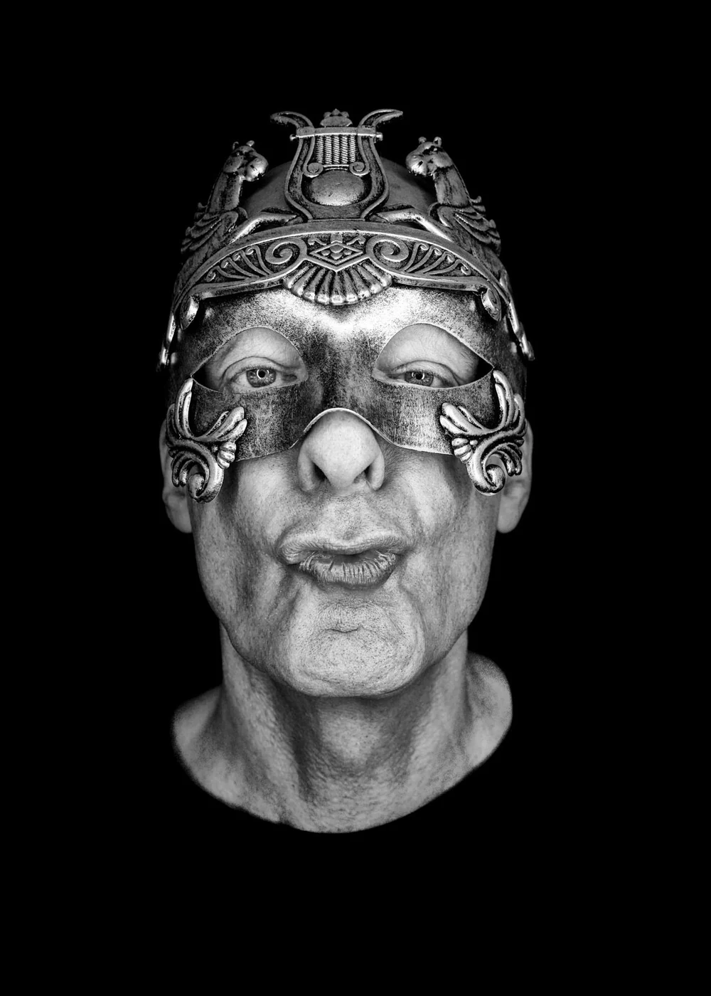 Black and white photo of an elderly person wearing a decorative metallic mask and a metallic helmet, against a black background.