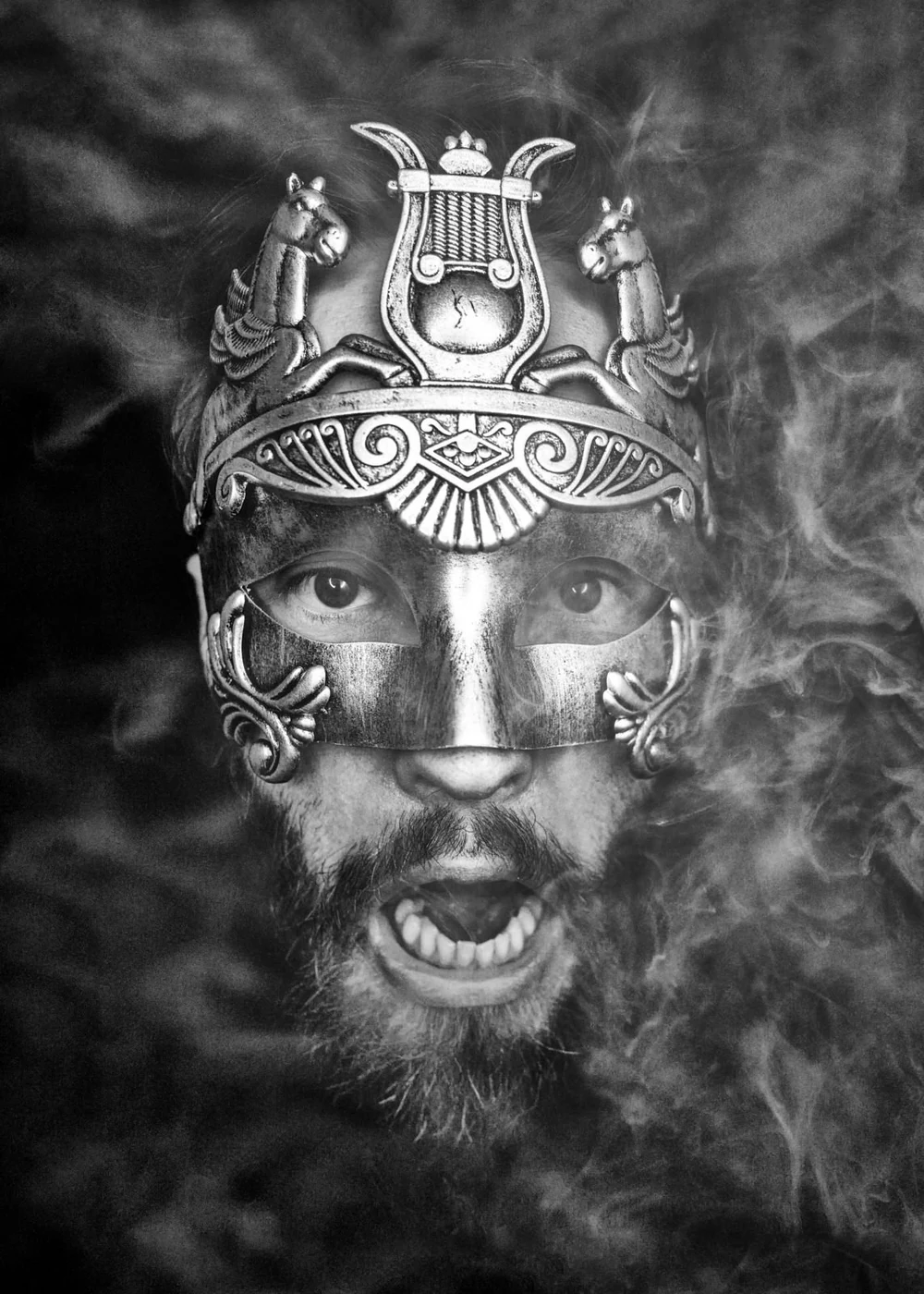 A person with a beard and mustache wearing a detailed, ornate metallic mask covering the eyes and nose, and a decorative headpiece with horse figures, surrounded by swirling smoke.