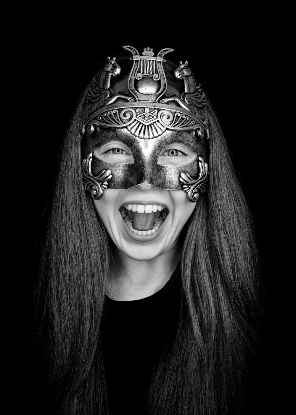 A woman with long hair wearing an ornate metallic mask that covers her eyes and forehead, with her mouth open in a joyful expression against a black background.