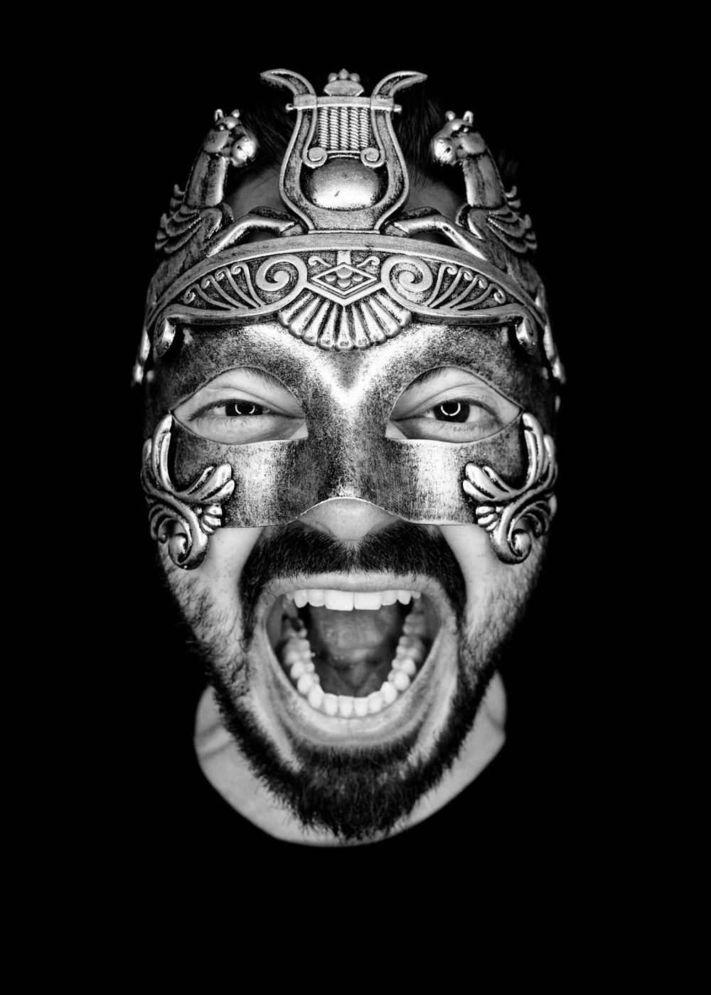 A man with a beard and mustache wearing a detailed metallic mask with a dragon design, open-mouthed expression, in black and white.