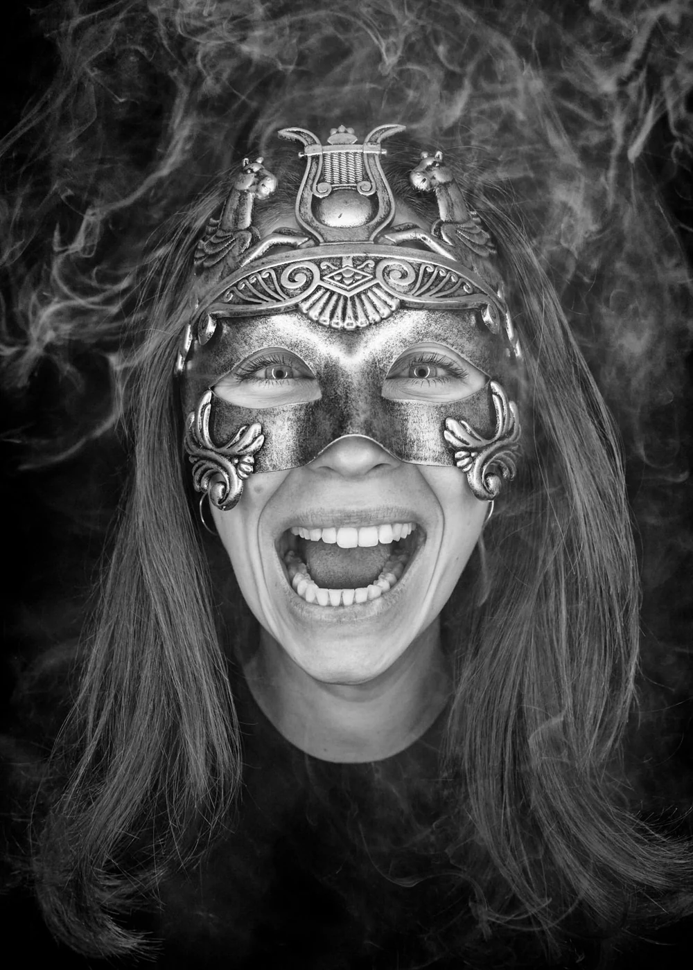 A woman wearing an ornate metallic mask with animal designs, smiling widely with her mouth open, surrounded by swirling smoke.